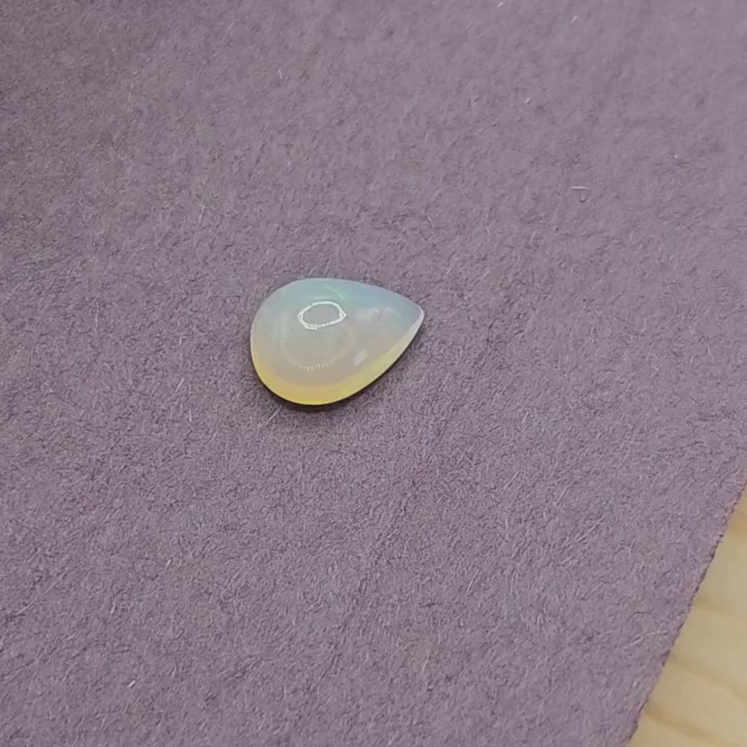 Real White Ethiopian Opal in pear shape, showcasing luminescence on purple fabric background.