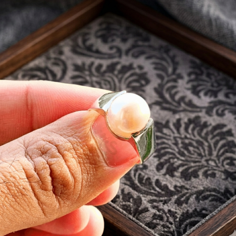 Hand holding a ring with a pearl and green stone against a patterned background
