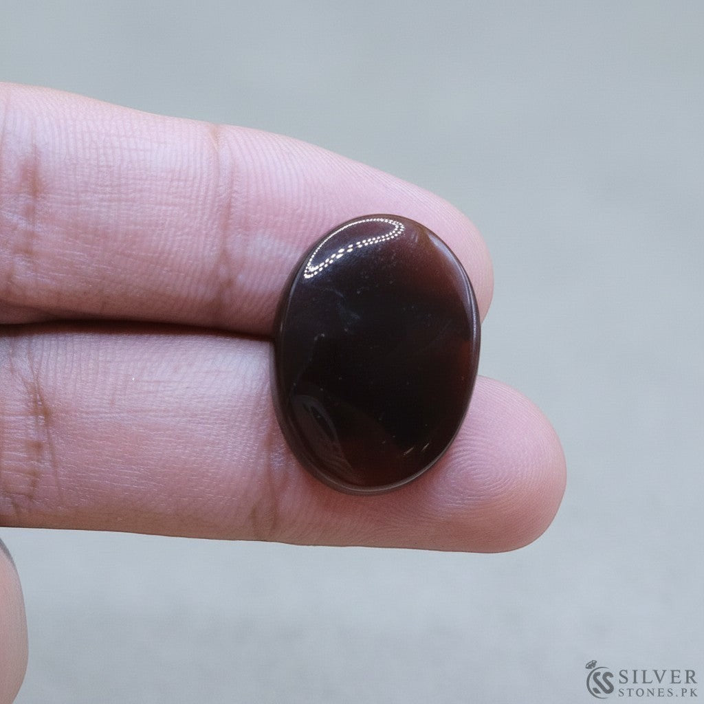 SilverStones.Pk oval-cut real Sulemani Aqeeq stone, deep brown with reddish-orange banding, held by a thumb and forefinger.